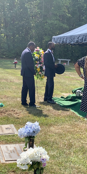 Two men in suits stand near a floral arrangement at a graveside service.