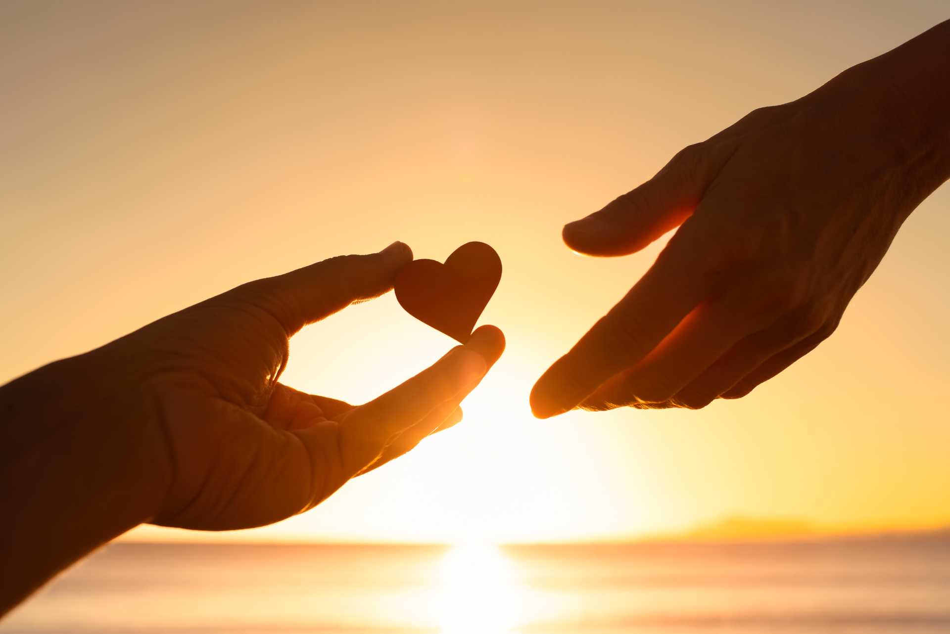 Two hands reaching toward a small heart at sunset over the water