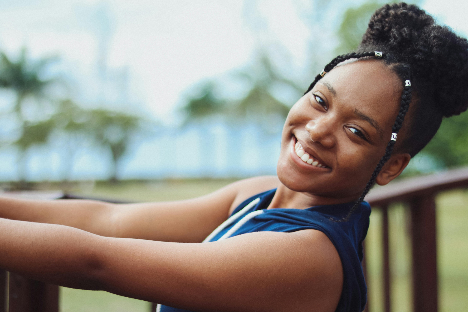 Smiling person leaning on a wooden railing outdoors, with a blurred beach background.