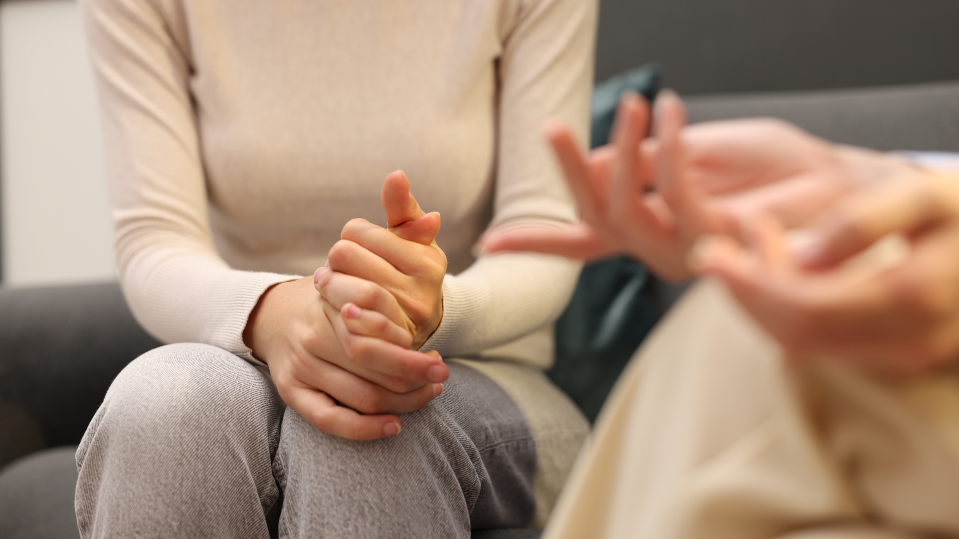 Two people seated in conversation, one clasping hands, in a softly lit room.