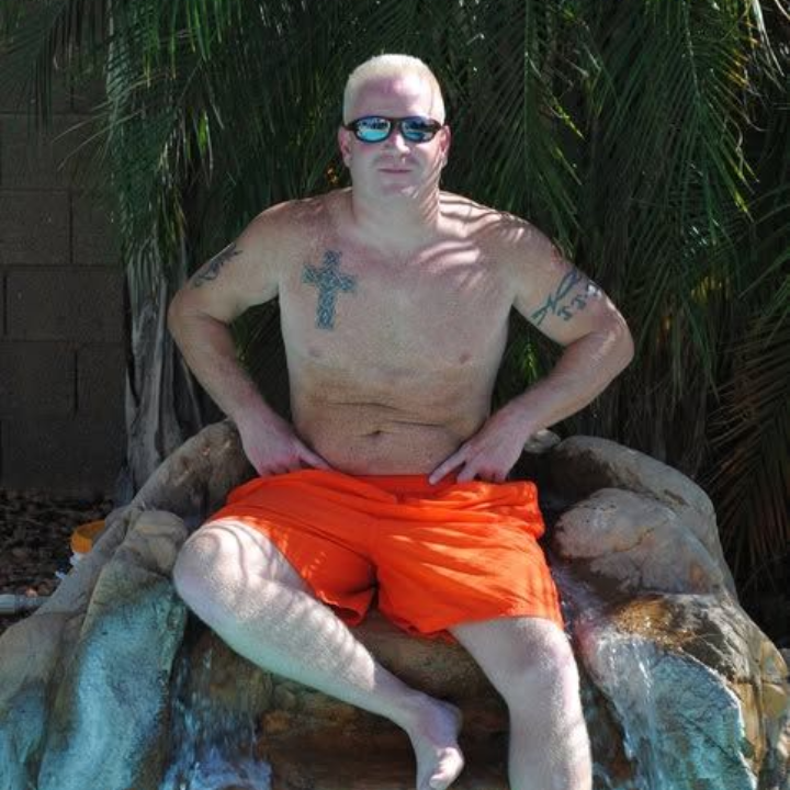 Man sitting on a rock formation in orange shorts and sunglasses outdoors, with tattooed chest and arms.