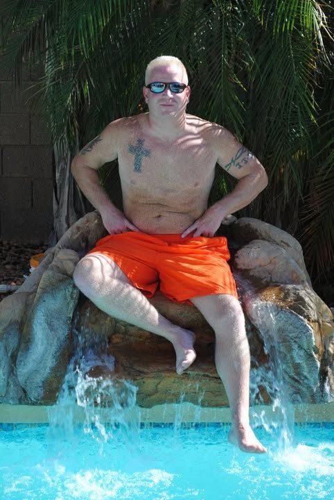 Man in orange shorts and sunglasses sitting on a rock beside a swimming pool with waterfall backdrop