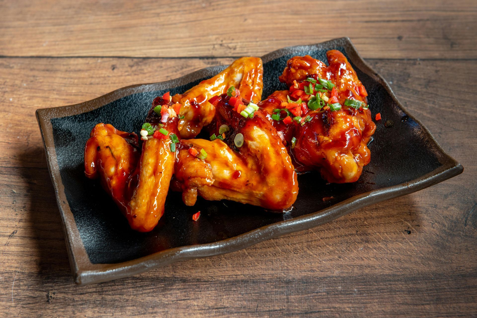Glazed chicken wings on a black platter, topped with chopped herbs and sauce on a wooden table