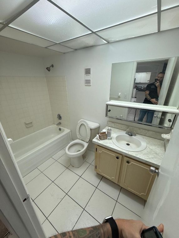 Small beige bathroom with tub, toilet, vanity, mirror, and a person reflected in the mirror