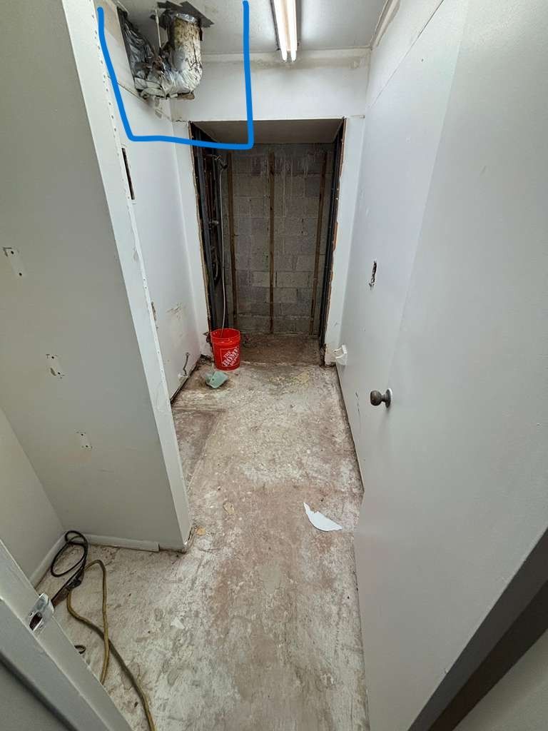 Narrow utility closet under renovation with exposed wall cavity, red bucket, and dusty floor