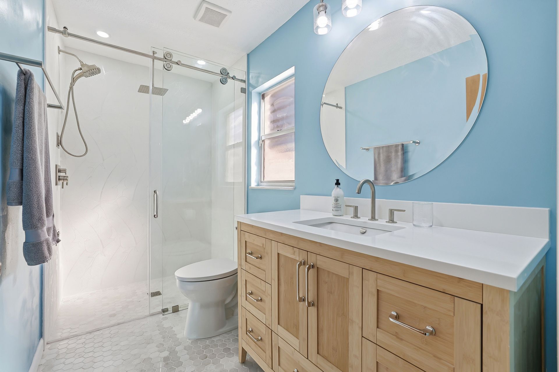 Bright bathroom with glass shower, white toilet, and wood vanity with round mirror on blue wall