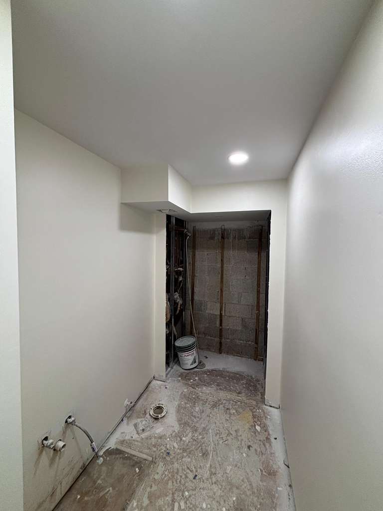 Narrow unfinished hallway with white walls, exposed floor, and a lit ceiling light leading to a dark doorway.