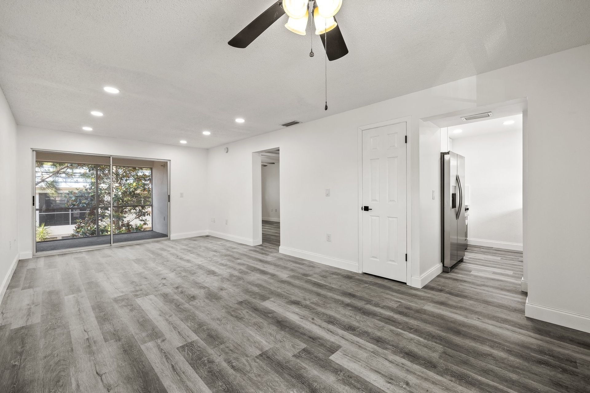 Bright empty living room with gray floors, white walls, ceiling fan, and large sliding glass doors