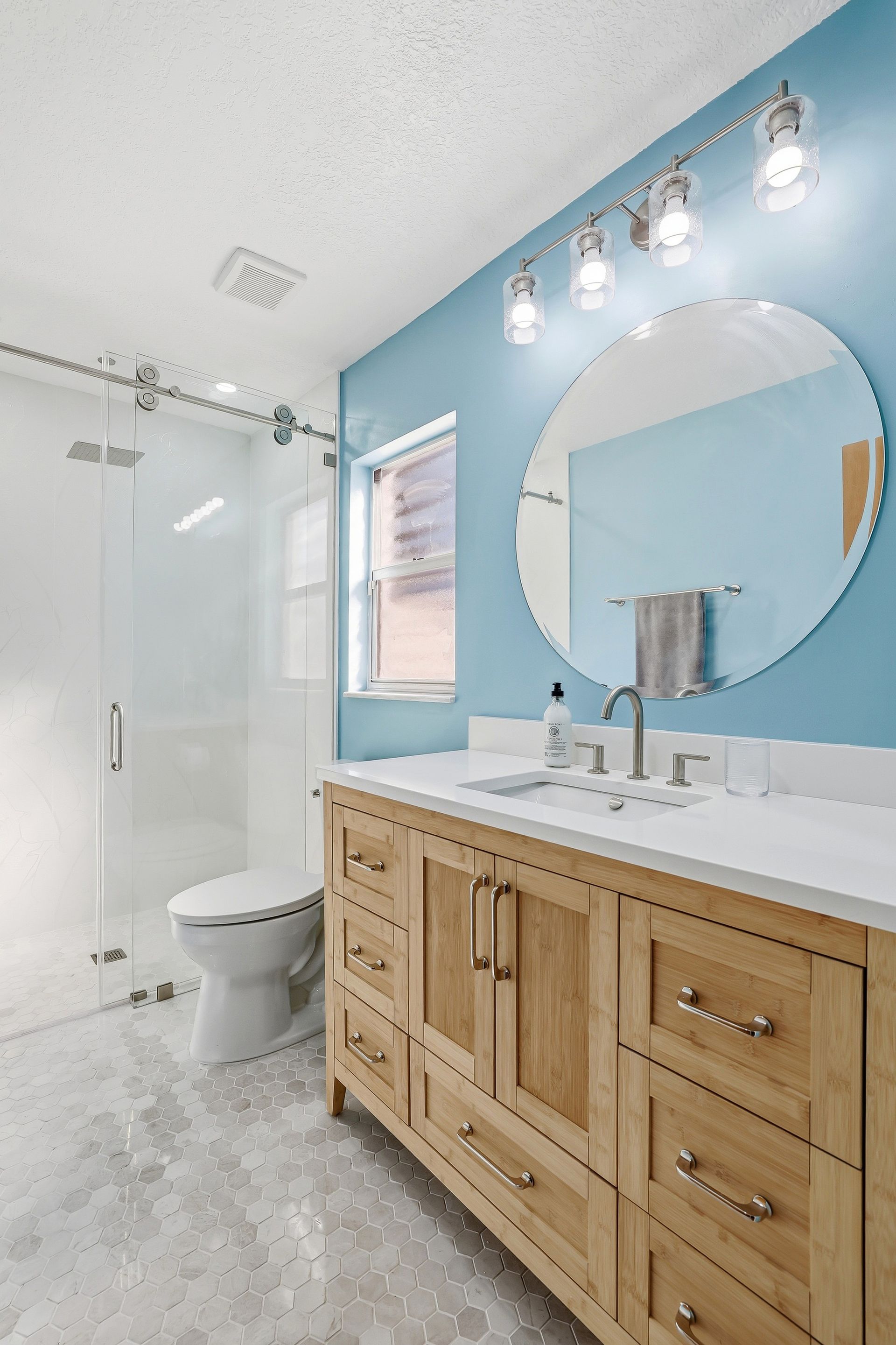 Bright blue bathroom with wood vanity, round mirror, and glass shower door