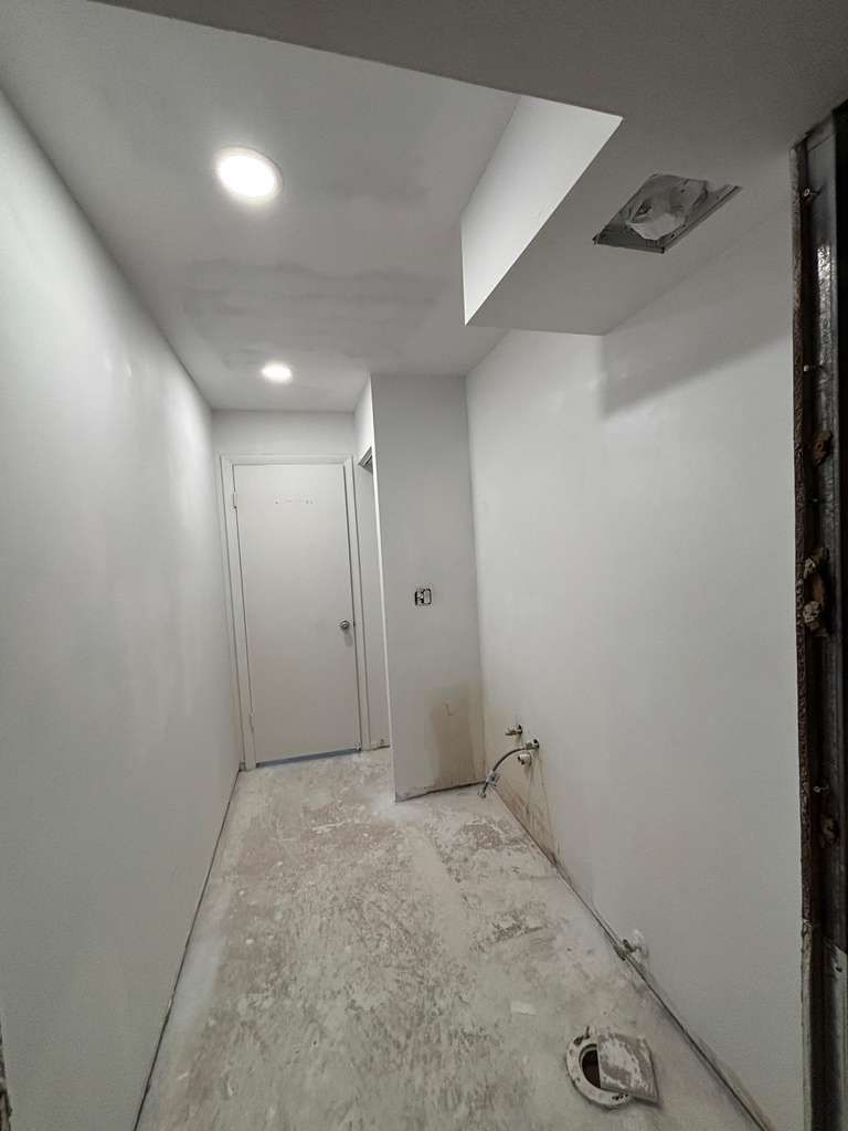 Narrow white hallway under construction with recessed lights, bare floor, and a door at the end.
