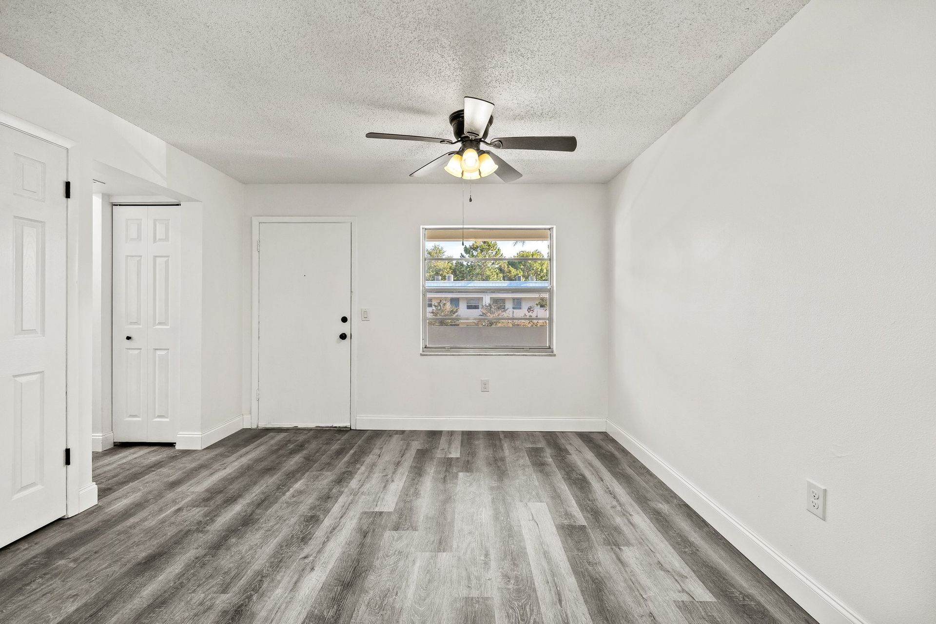 Empty white bedroom with gray carpet, ceiling fan, closet doors, and a window with blinds.