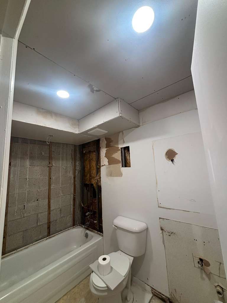 Small bathroom with toilet, bathtub, sink area, and ceiling lights; some wall damage visible.