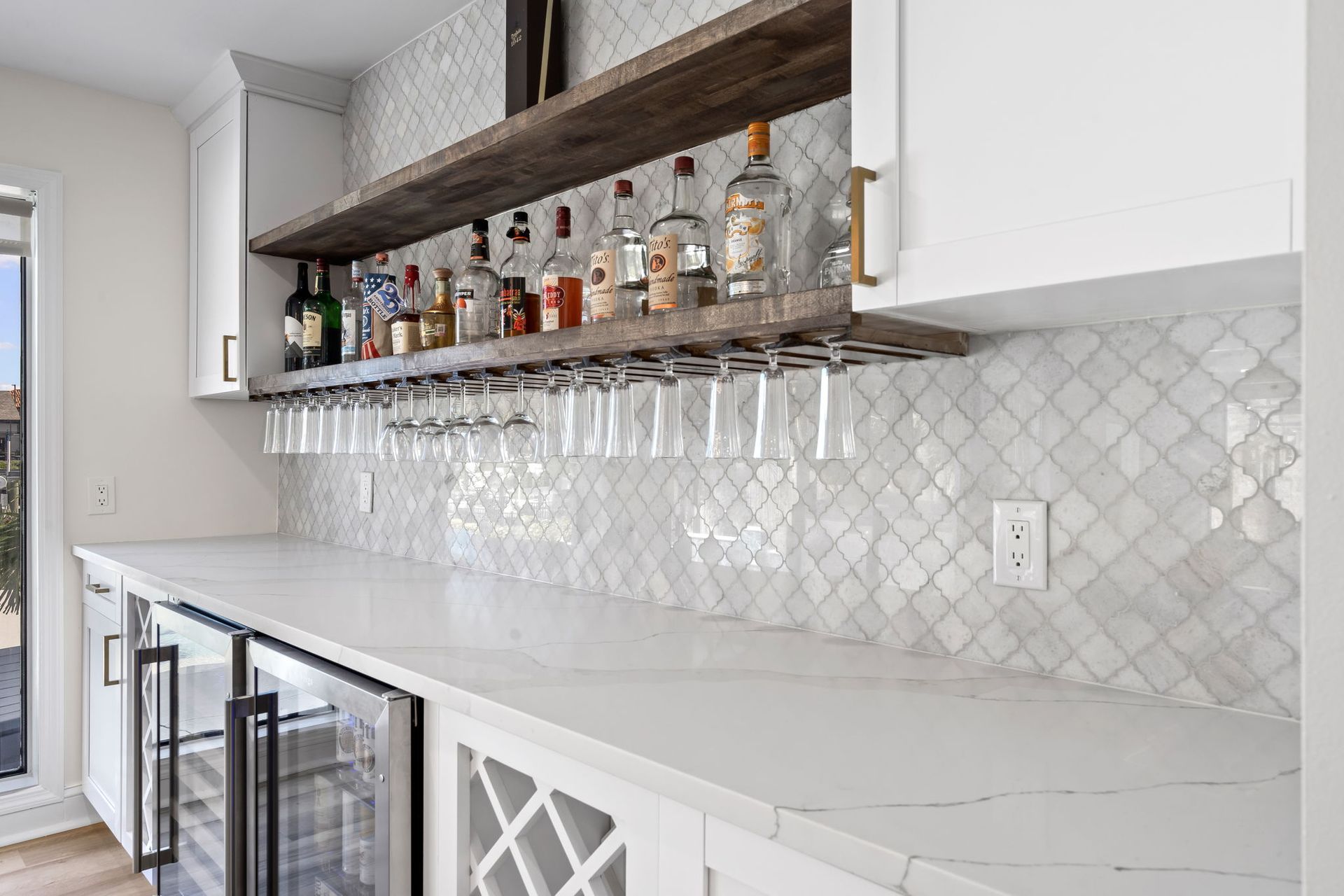 Modern home bar with glass shelves, hanging stemware, bottles, and white tiled backsplash