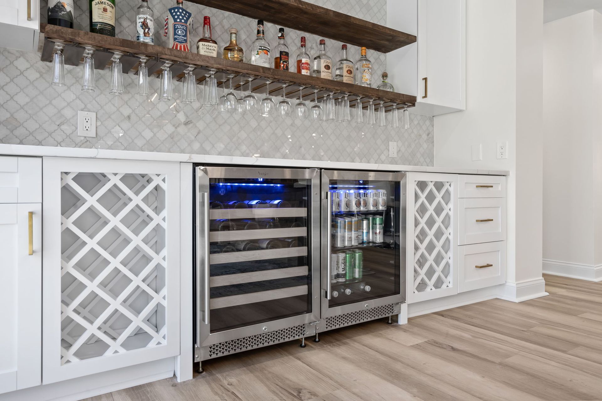 Built-in home bar with wine fridge, beverage cooler, hanging stemware, and white lattice cabinets
