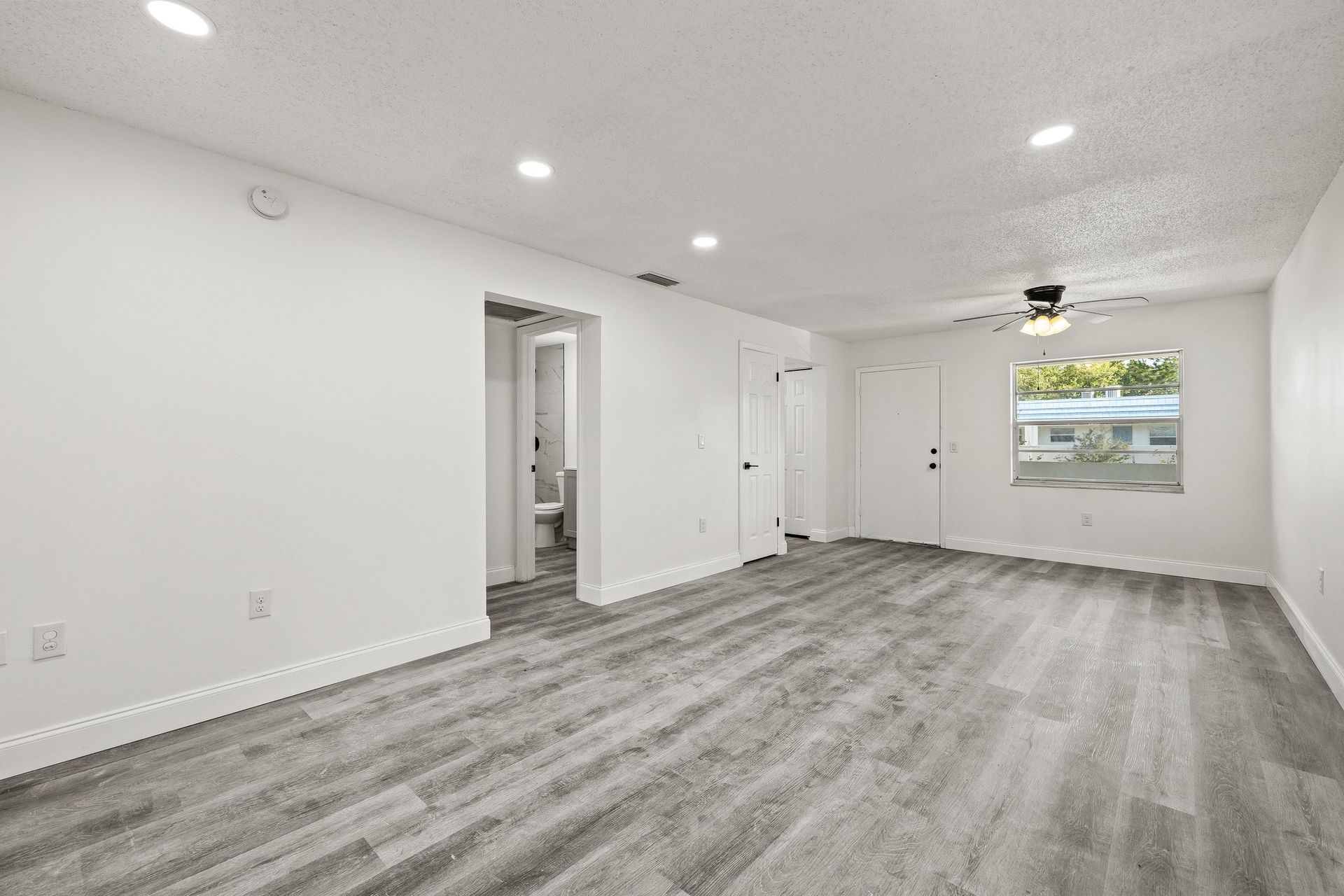Empty bright room with gray carpet, white walls, recessed lights, and a window on the far wall