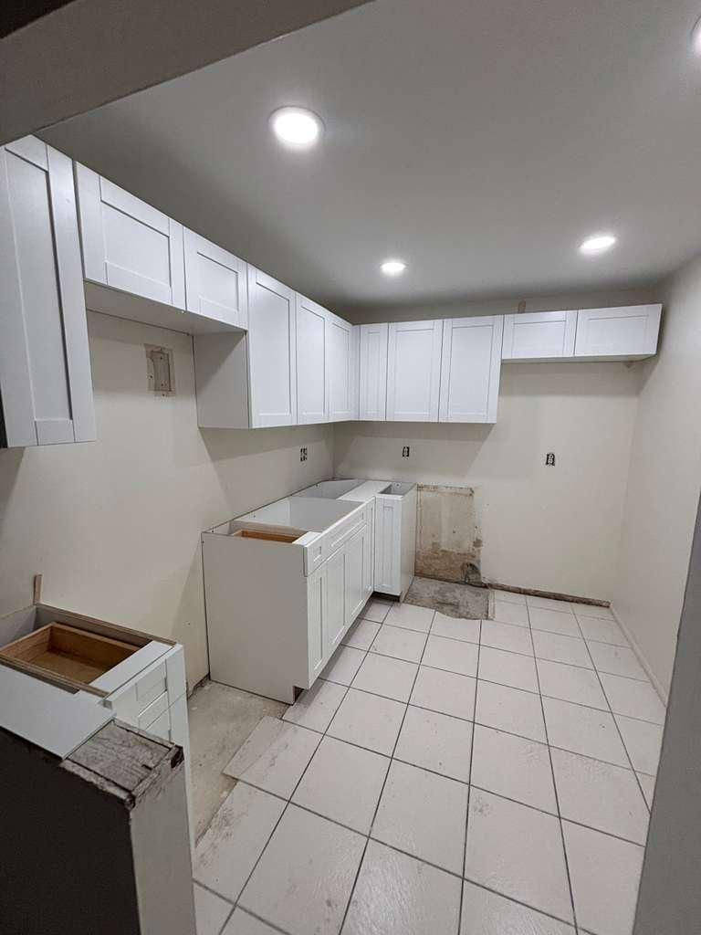 Empty white kitchen with tiled floor, cabinets, sink, and recessed ceiling lights.