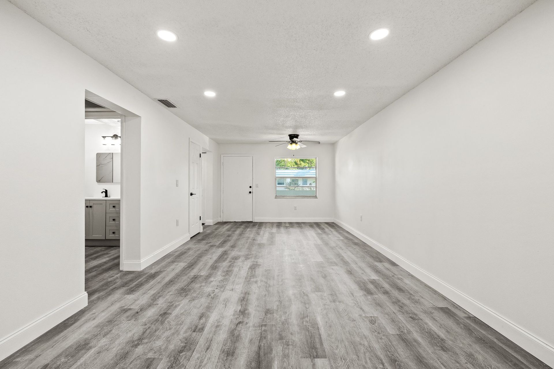 Bright empty living room with gray wood floors, white walls, recessed lights, and a view into the kitchen.