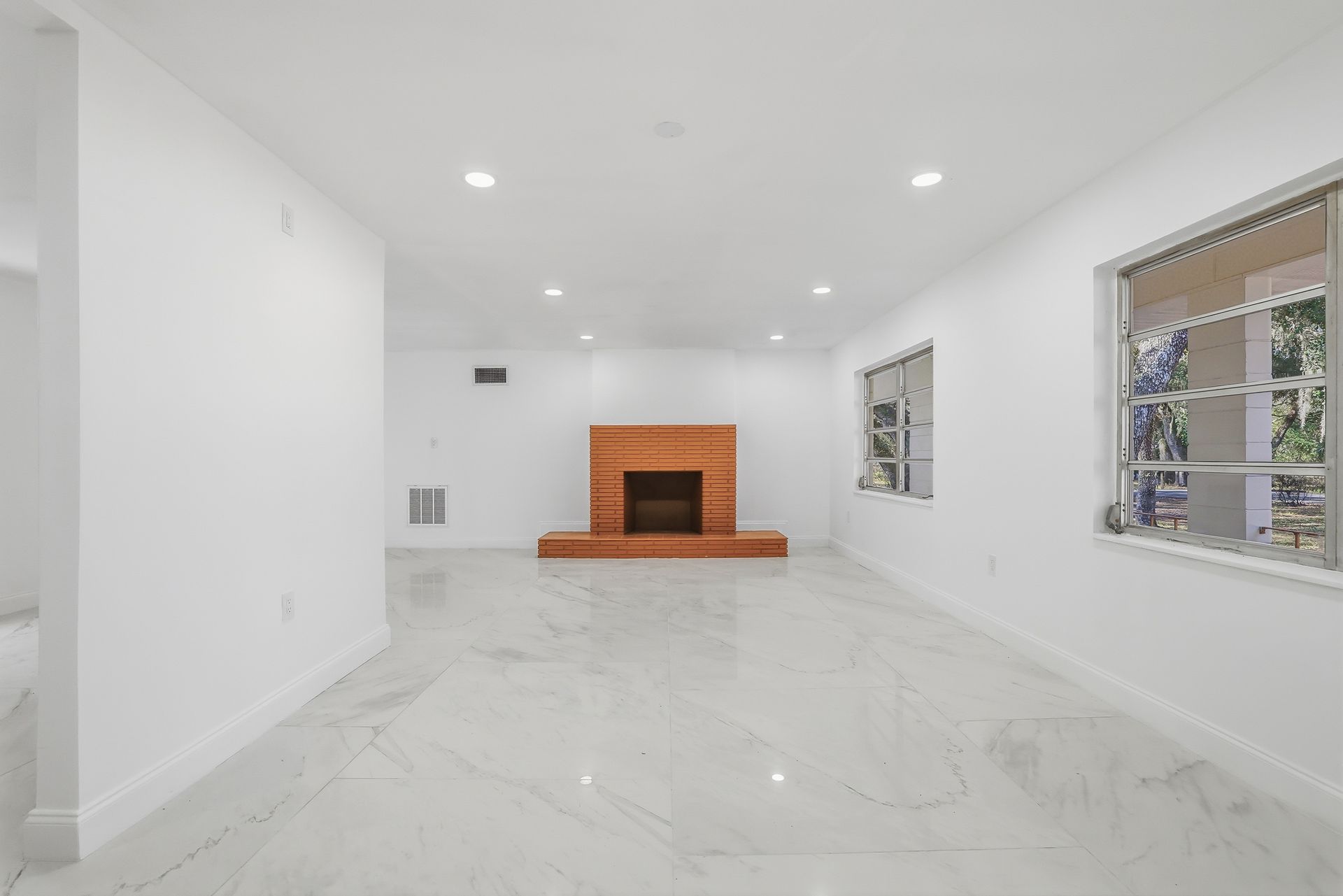 Bright empty living room with white tile floors, recessed lights, and a central brick fireplace