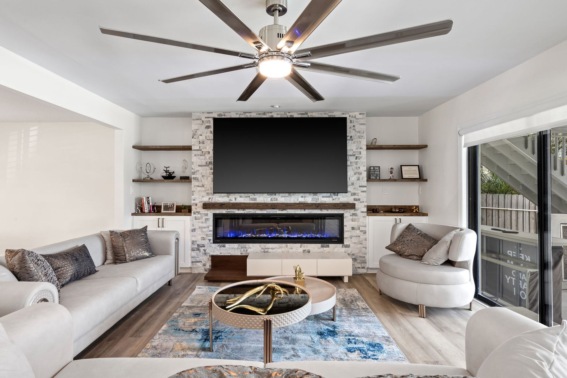 Bright living room with white furniture, stone fireplace, wall-mounted TV, and a ceiling fan