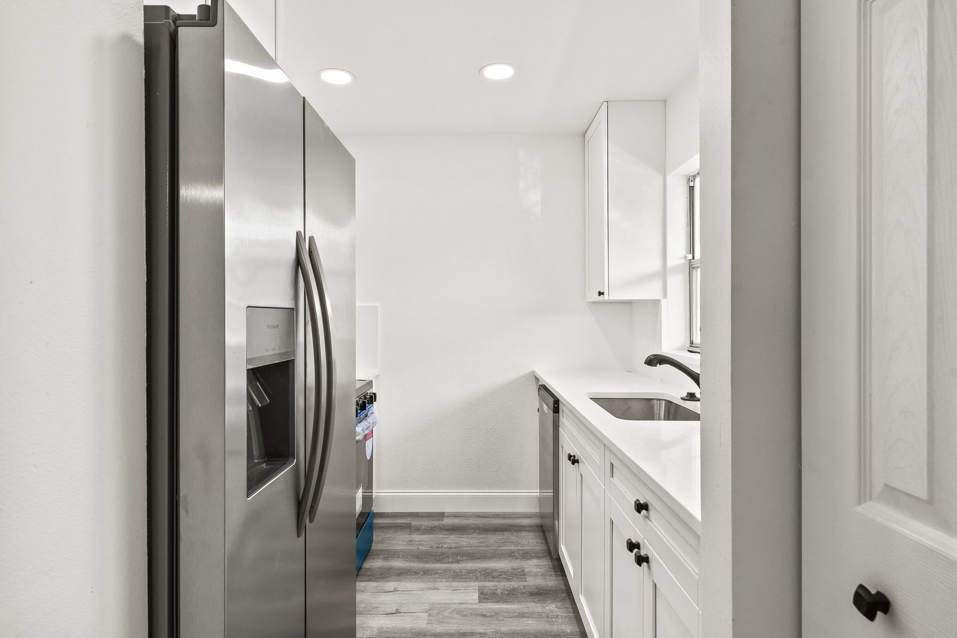 Bright white galley kitchen with stainless fridge, white cabinets, and gray wood floor