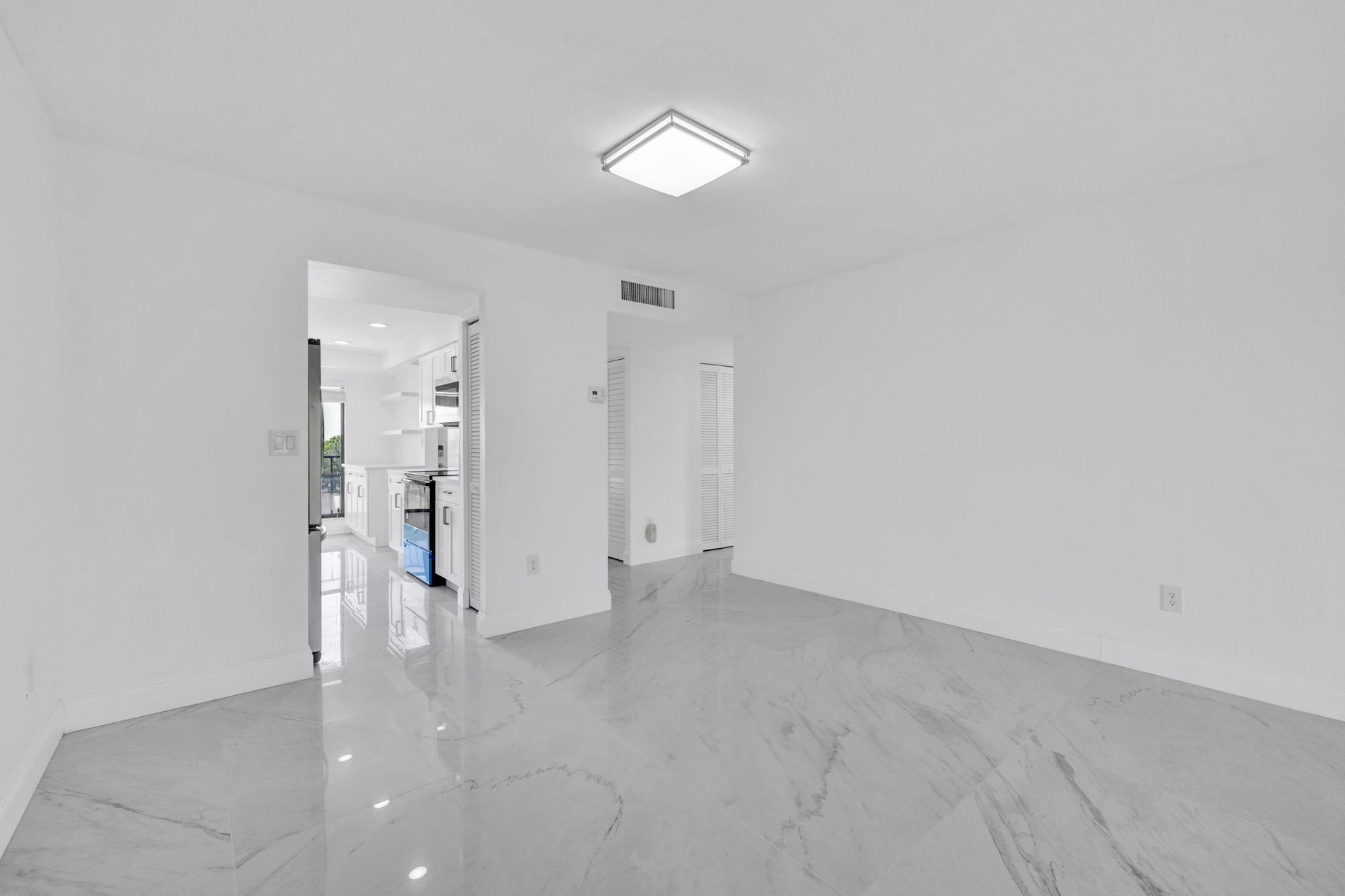 Empty white room with glossy marble floor, ceiling light, and doorway to a kitchen area