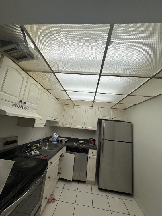 Small kitchen with white cabinets, tile floor, a stainless fridge, and fluorescent ceiling panels.