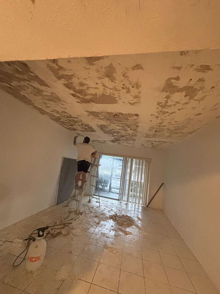 Worker scraping peeled ceiling plaster in a room with debris-covered floor and sliding glass doors.