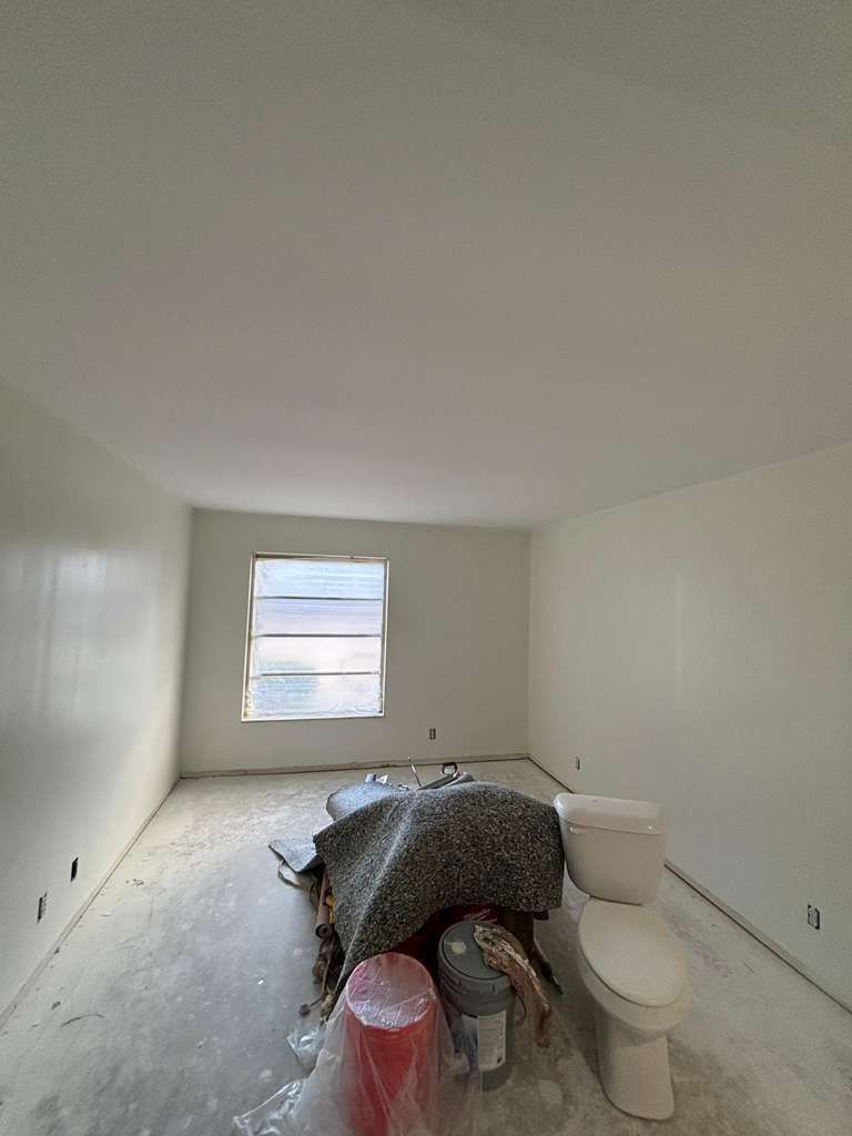 Unfinished white room with a window, toilet, and covered furniture during renovation.