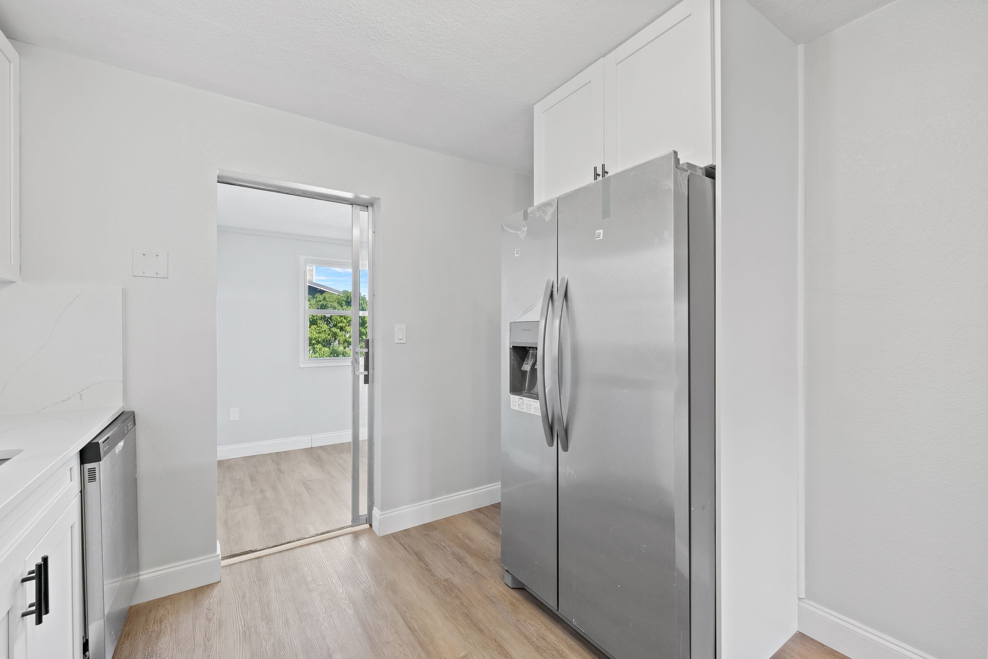 Bright white kitchen with stainless steel refrigerator and a doorway to a sunlit room.
