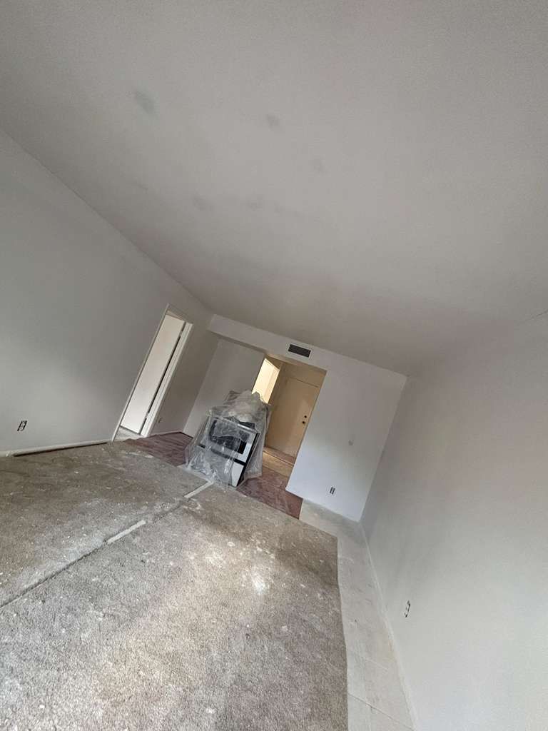 Empty white room under renovation with unfinished floor, open door, and a chair near the back.