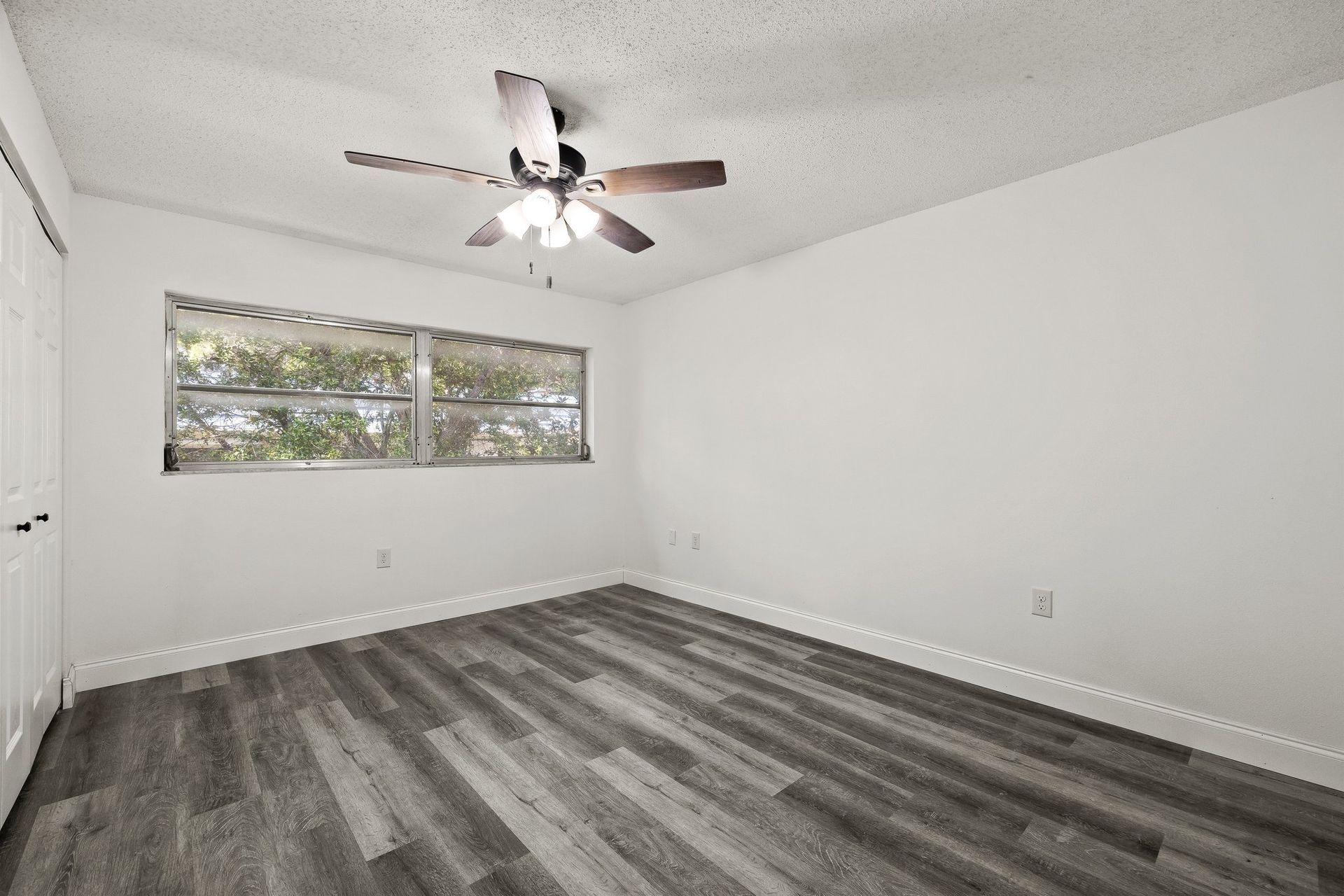 Empty white bedroom with gray wood-look flooring, ceiling fan, and a window with blinds
