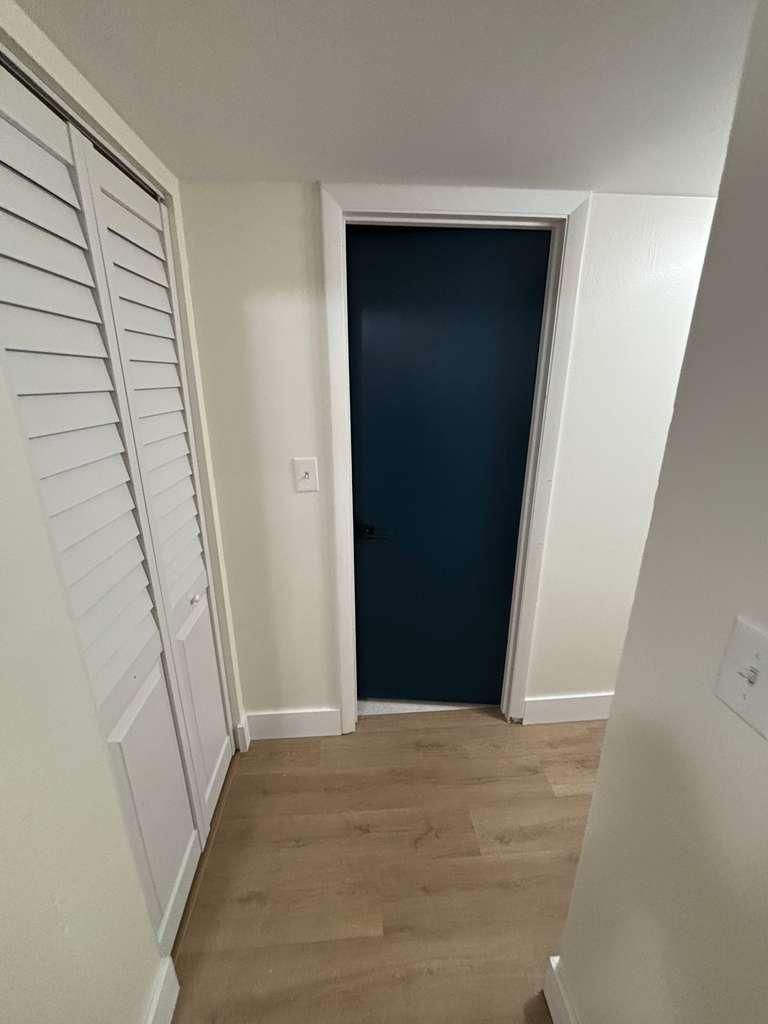 Narrow hallway with wood floor and white walls, featuring a dark blue open doorway ahead.