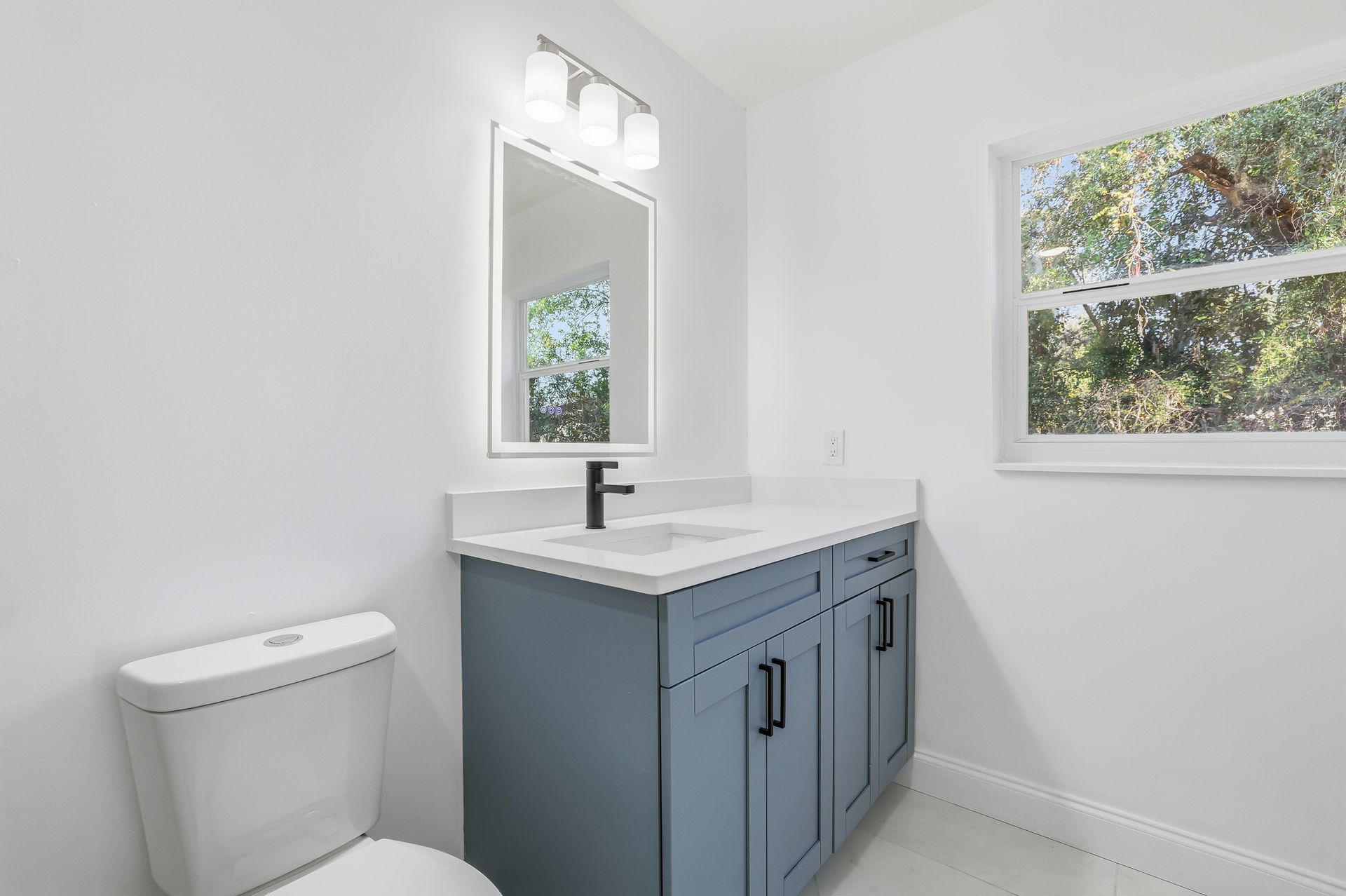Bright bathroom with blue vanity, white sink, mirror, toilet, and window.