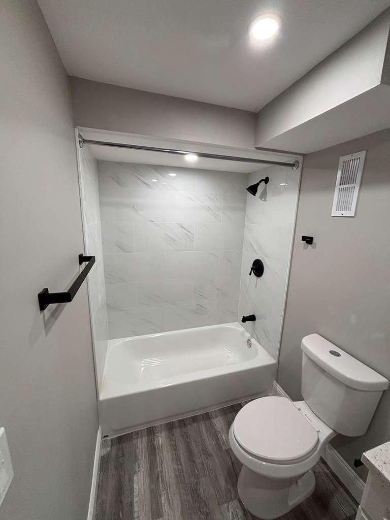 Small modern bathroom with a bathtub, toilet, grab bars, and gray walls.