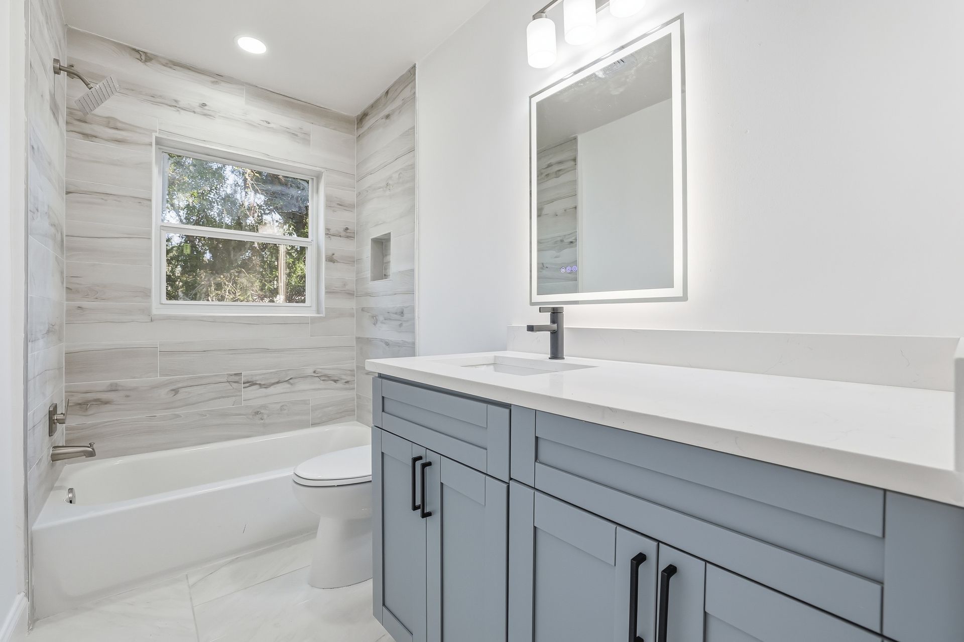 Bright bathroom with gray vanity, white countertop, mirror, window, and tiled bathtub surround