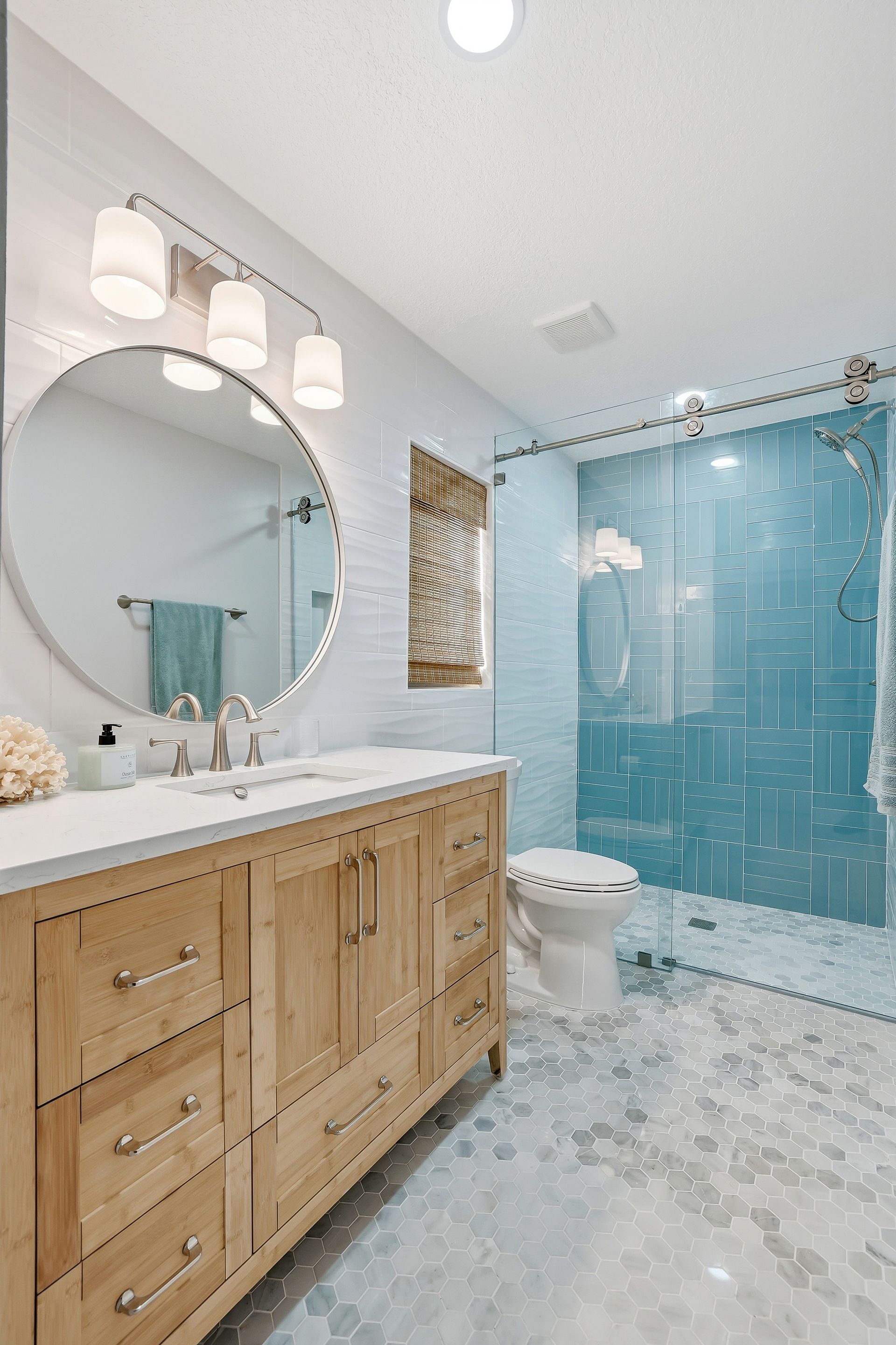 Bright bathroom with wood vanity, round mirror, marble floor, and blue tiled shower with glass door