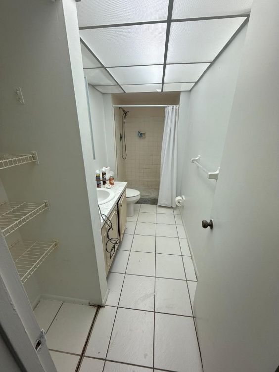Narrow white bathroom with tiled floor, sink vanity, wire shelves, and a shower curtain at the far end