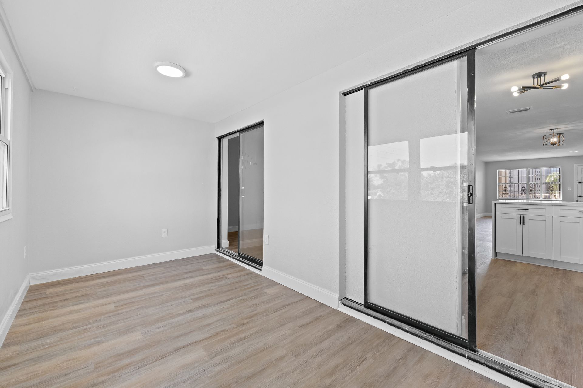 Empty white room with wood floor and sliding glass closet doors leading to a bright kitchen area.