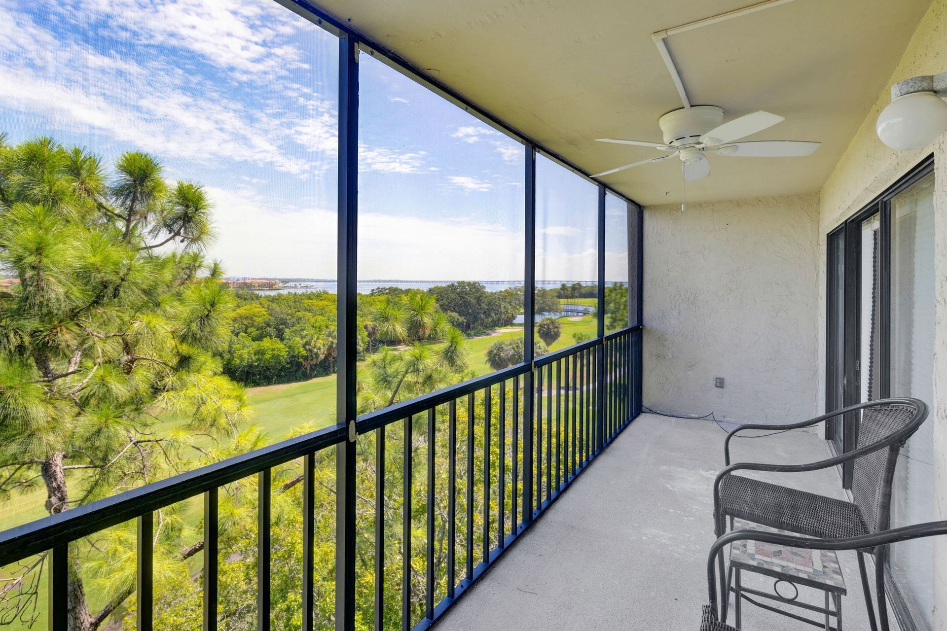 Covered balcony with black railings, wicker chairs, ceiling fan, and view of green trees and sky