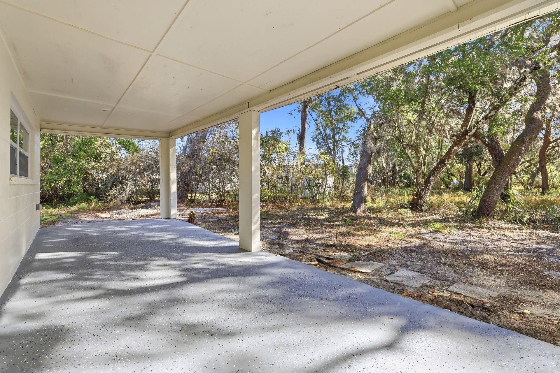 Covered concrete patio beside a white house, bordered by trees and brush.
