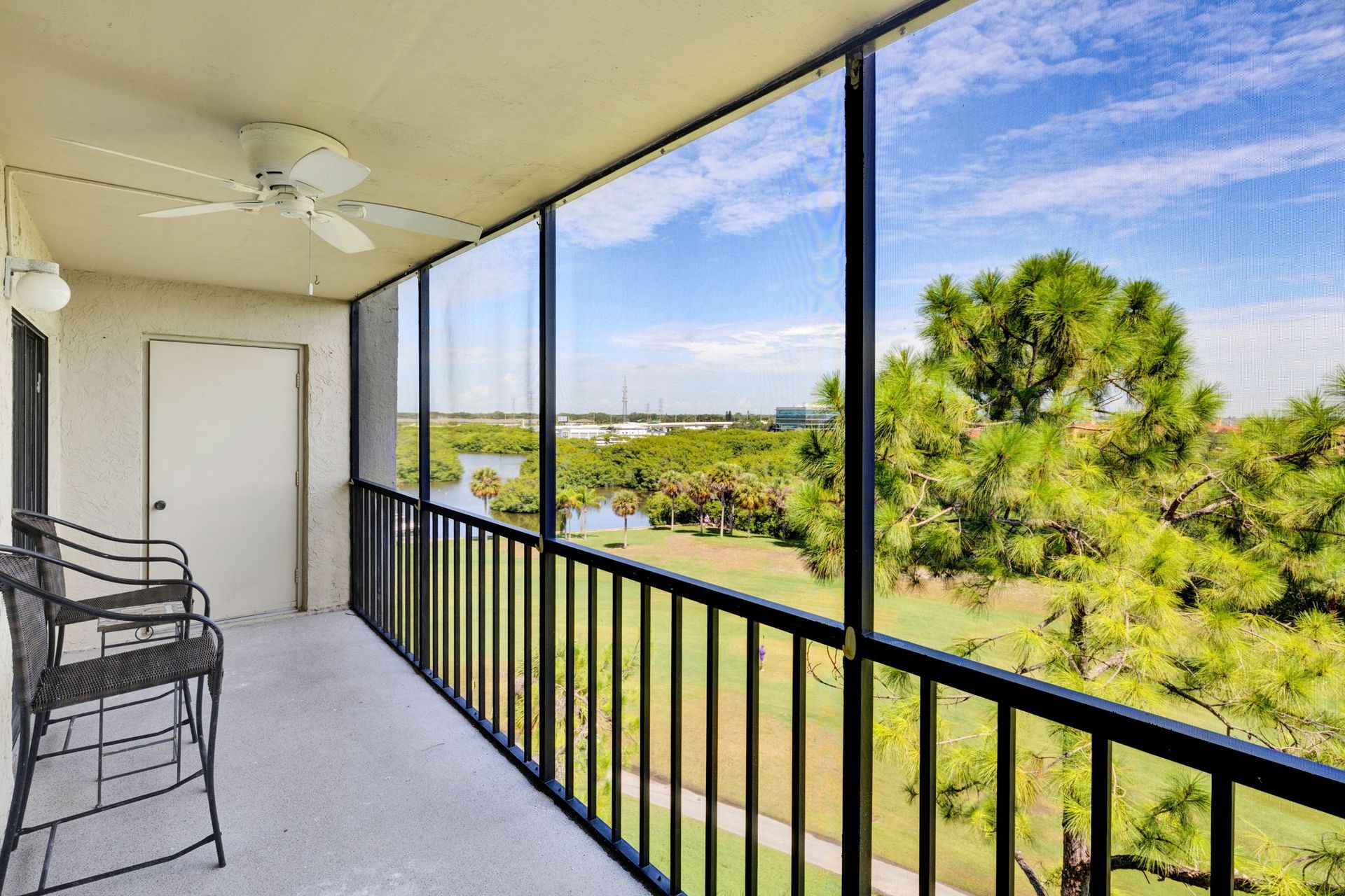 Screened balcony with black railing, chairs, and a view of green lawn and trees under a blue sky