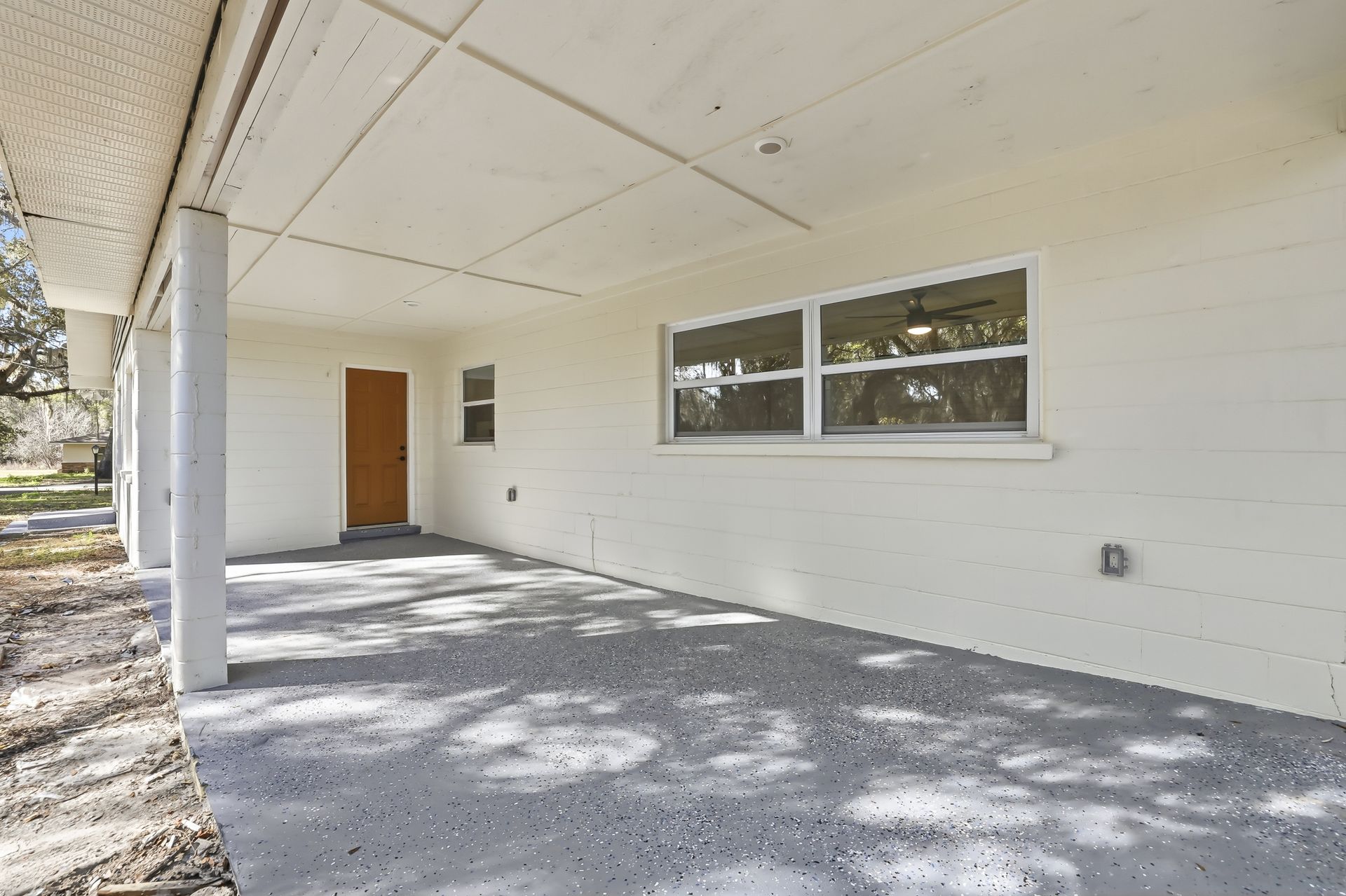 Covered concrete porch with white wall, brown door, and a large window in sunlight.