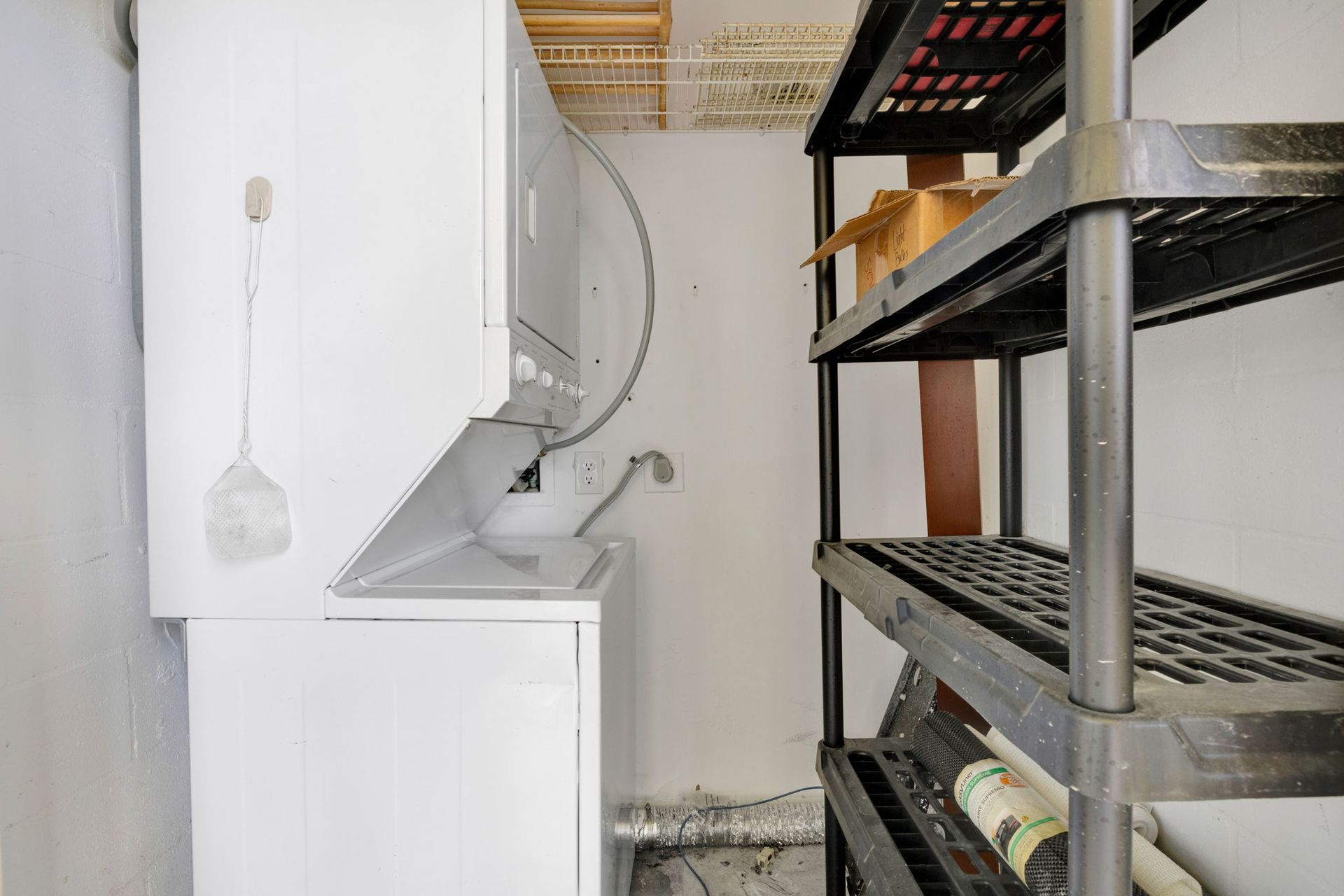Narrow utility room with white washer and dryer, metal shelving, and cleaning tools.
