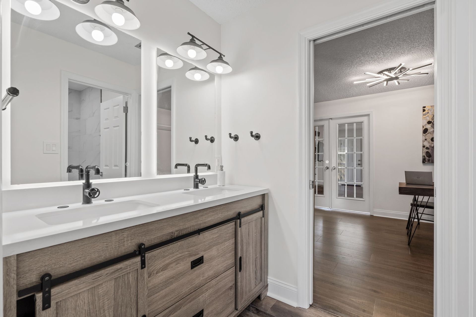 Bright bathroom with double vanity, mirrors, and pendant lights, opening to a room with wood floors.