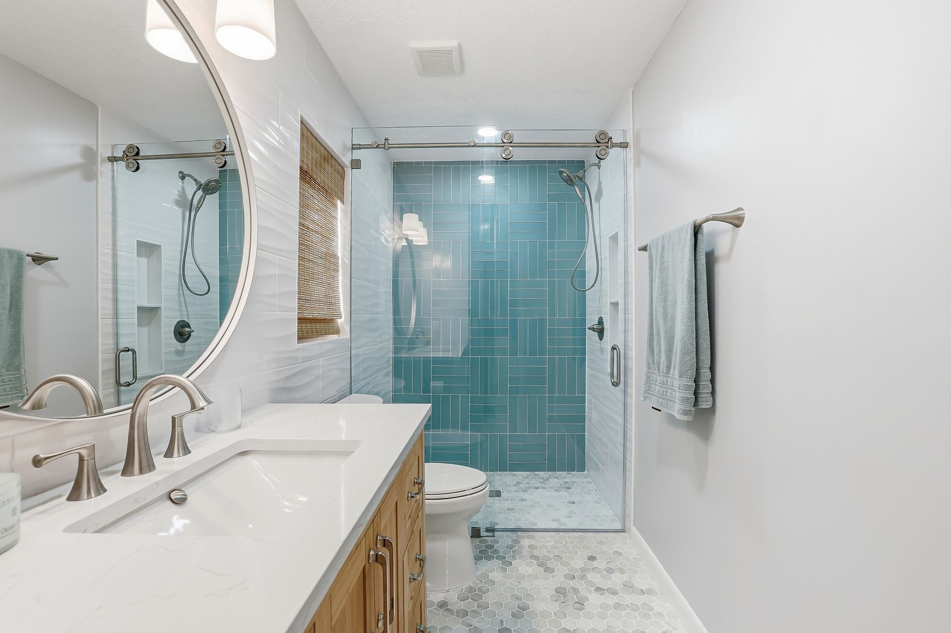 Bright bathroom with double sinks, white counters, wood vanity, and blue glass shower enclosure