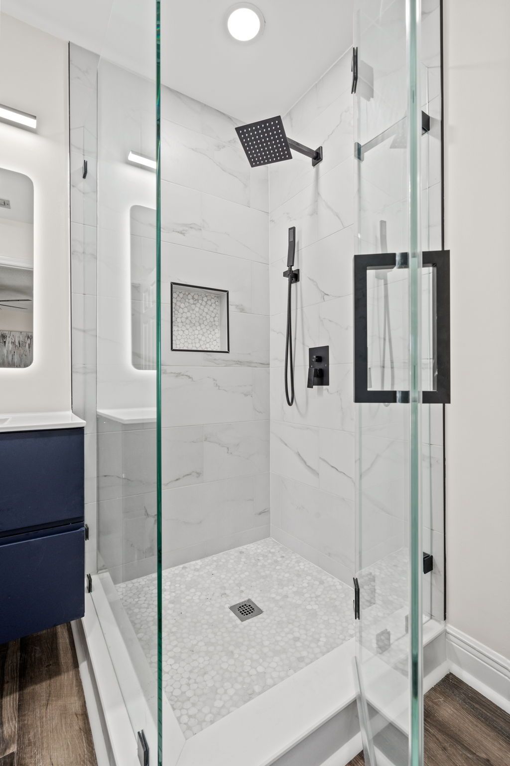 Modern glass shower with white tile, black fixtures, and a navy vanity visible nearby