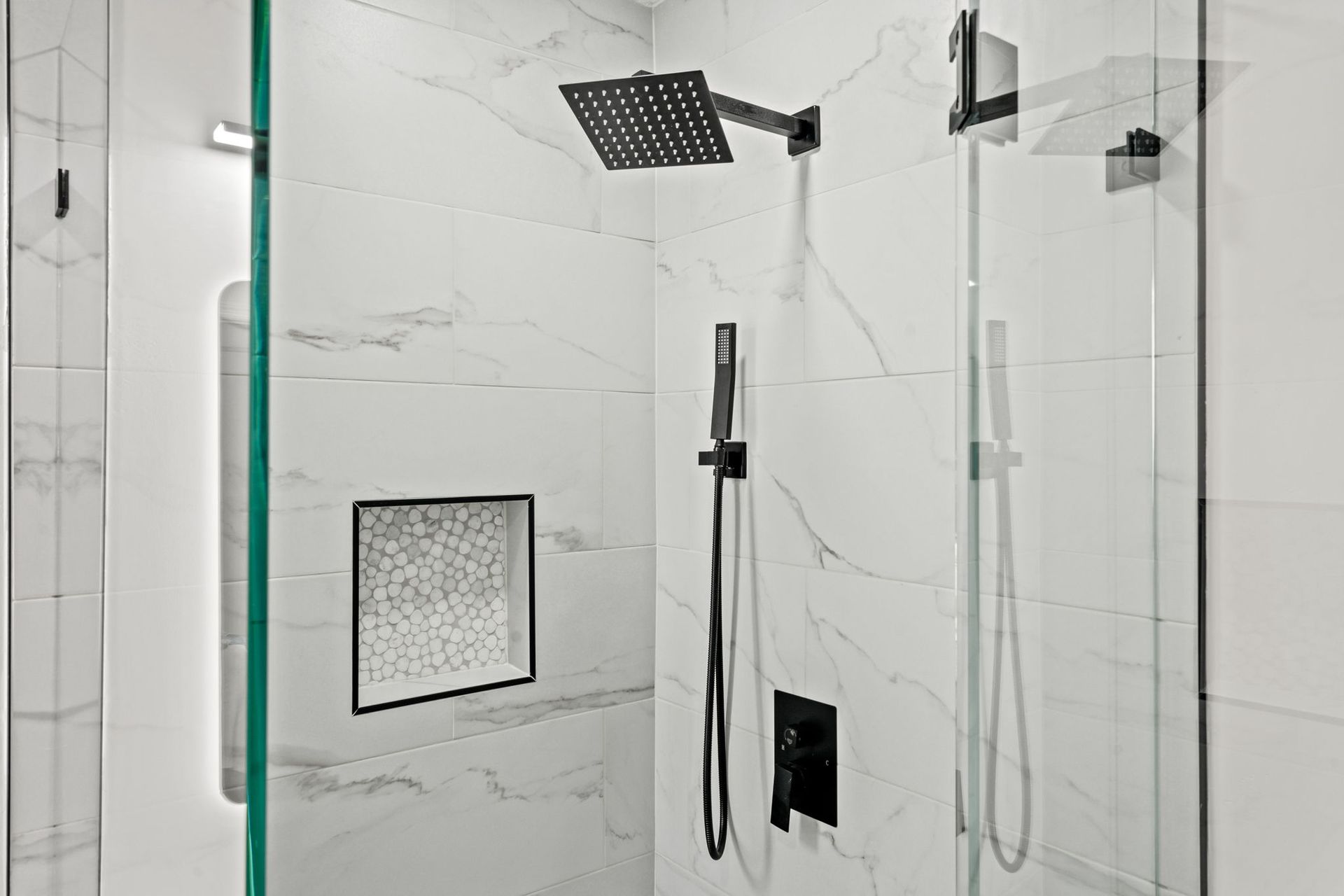 Modern white-tiled shower with glass doors, black fixtures, rainfall head, handheld wand, and niche shelf