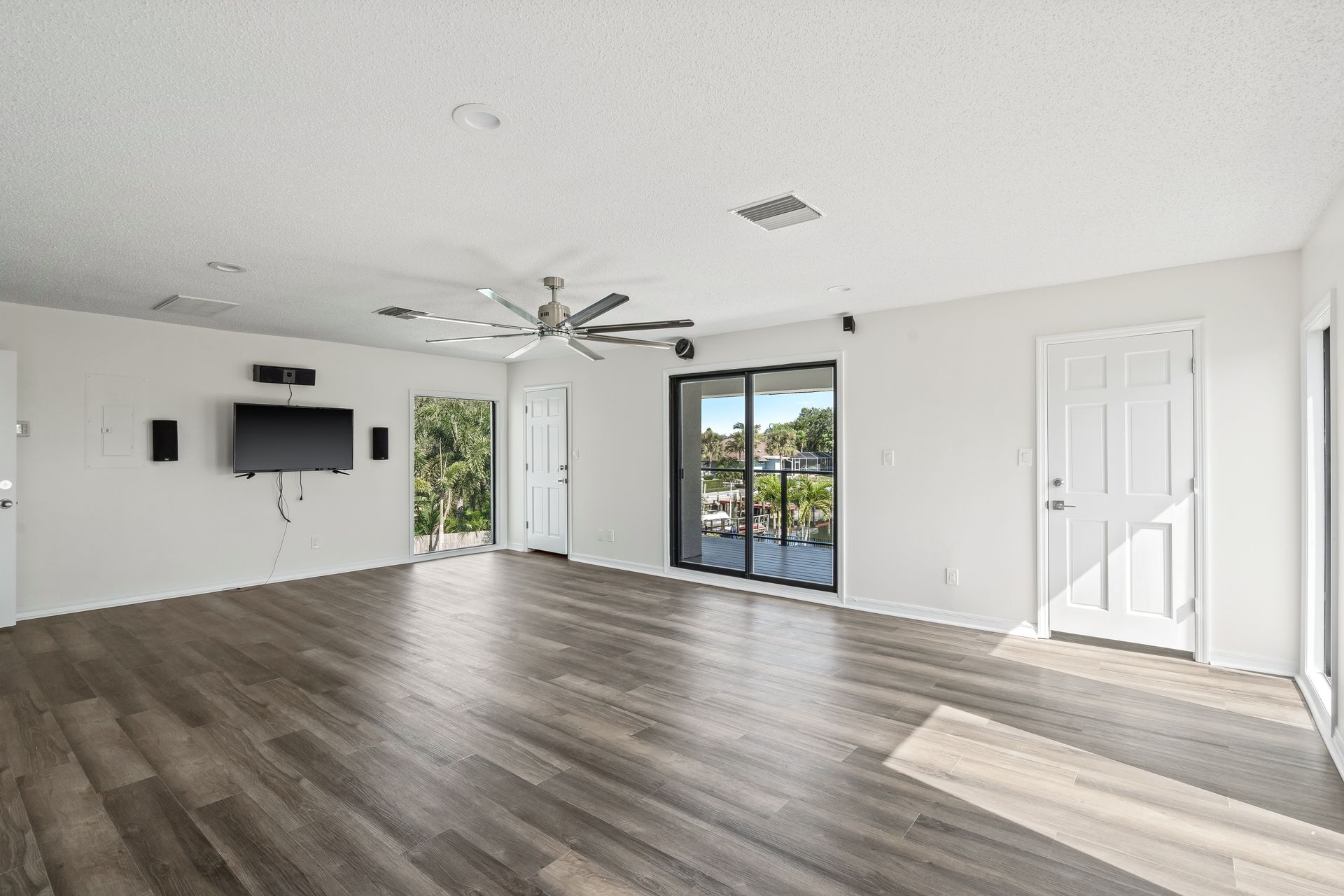 Empty modern living room with dark wood floors, white walls, TV mount, ceiling fan, and sliding glass door.