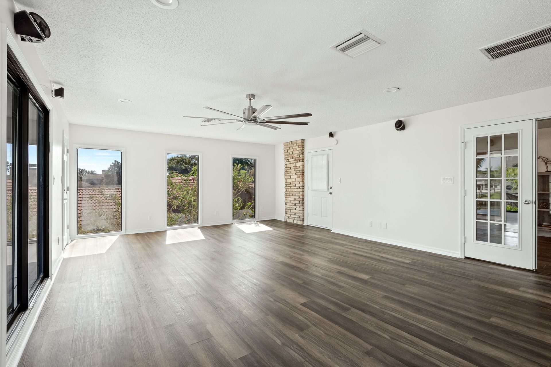 Empty modern living room with gray wood floors, white walls, large windows, and a ceiling fan.