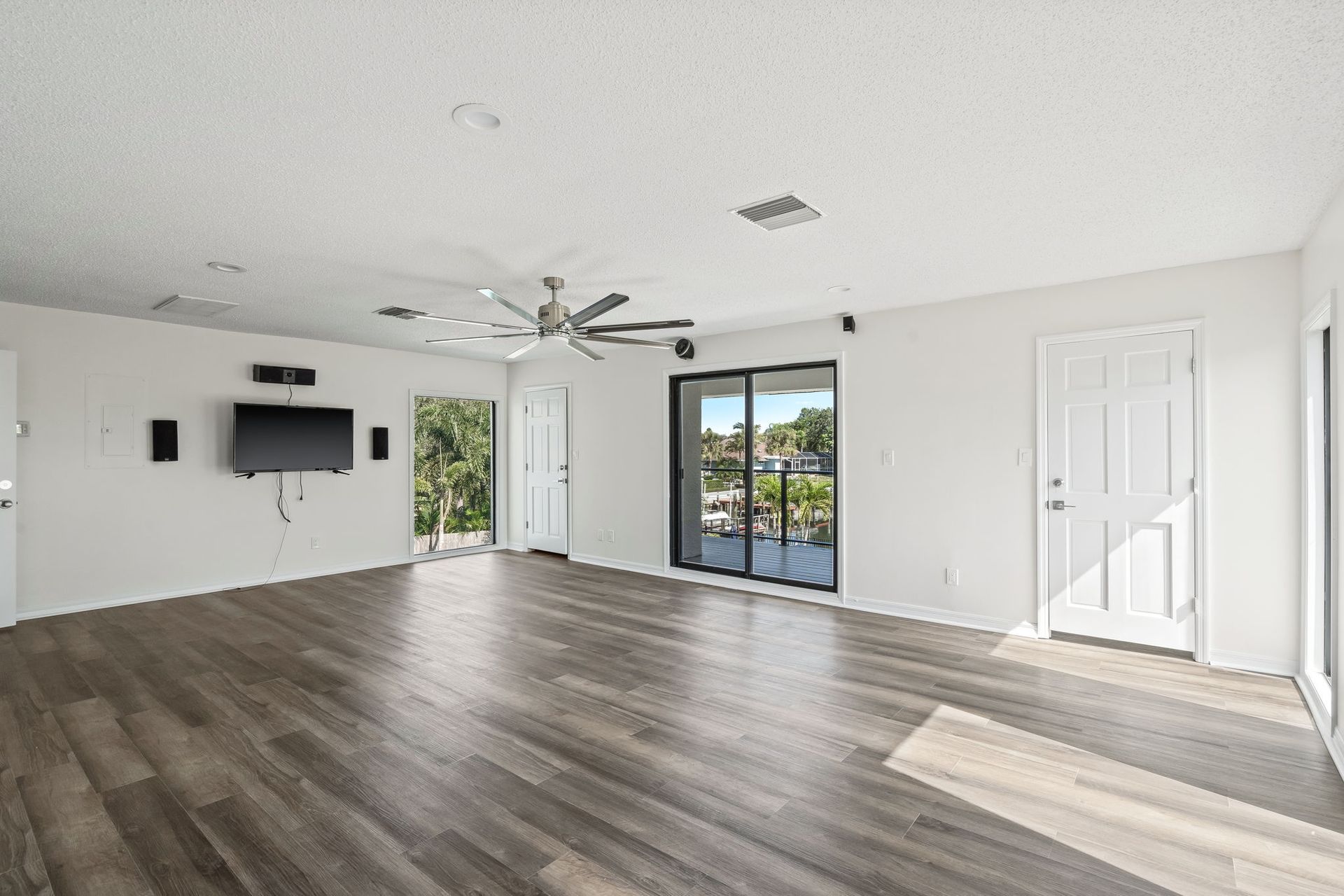 Spacious empty living room with dark wood floors, white walls, ceiling fan, TV mount, and sliding glass door